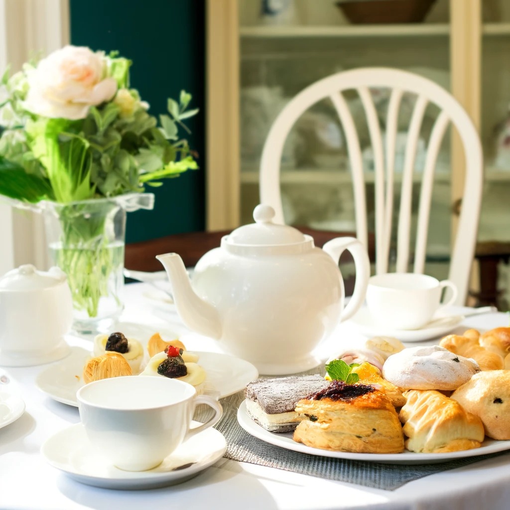 A beautifully set table for afternoon tea with pastries, scones, and a teapot on a white tablecloth, in a sunlit room with a vase of fresh flowers.